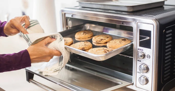 فردی در حال بیرون آوردن کوکی های پخته شده از درون فر. از میان لوازم پخت و پز برقی، آون توستر قابلیت گرم کردن و پختن انواع کیک و غذا را دارد.