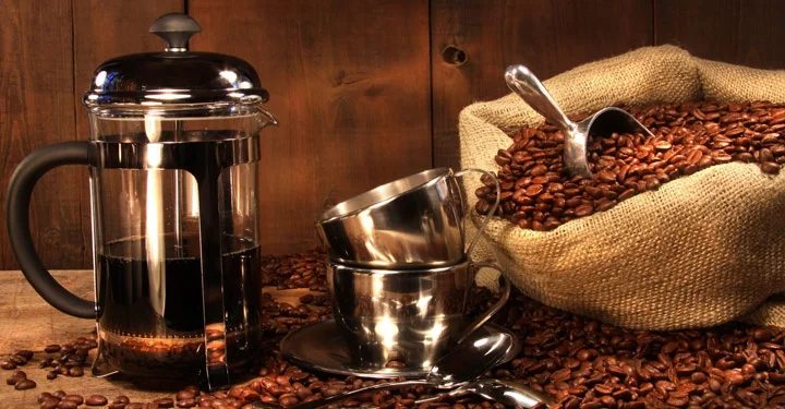 فرنچ پرسی که بر روی یک میز قهوه ای و کنار دانه های قهوه قرار گرفته است، این وسیله در دسته انواع قهوه ساز دستی قرار دارد.