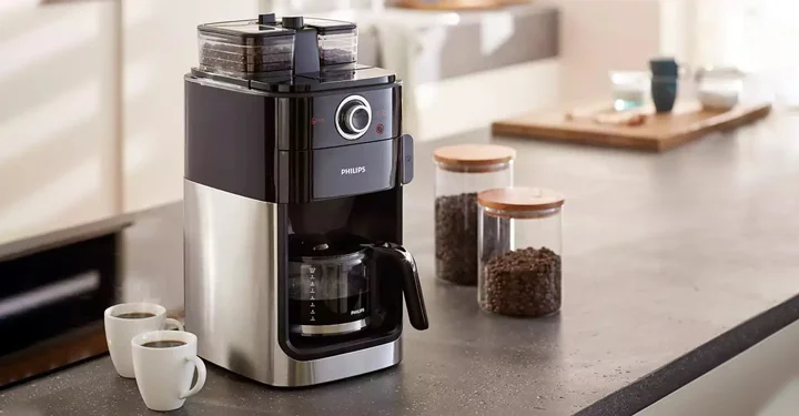 یک عدد قهوه ساز خانگی که دارای آسیاب قهوه می‌باشد و بر روی یک میز در کنار دو عدد فنجان سفید قرار گرفته است.