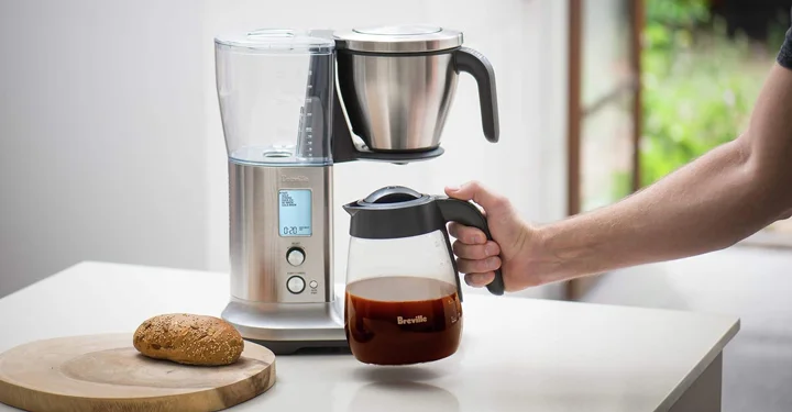 یک دستگاه قهوه ساز خانگی که بدنه آن از استیل و جنس کارافه آن شیشه است، این دستگاه بر روی یک سنگ قرار گرفته است و فردی کارافه را نگه داشته در کنار قهوه ساز یک تکه کیک قرار دارد.